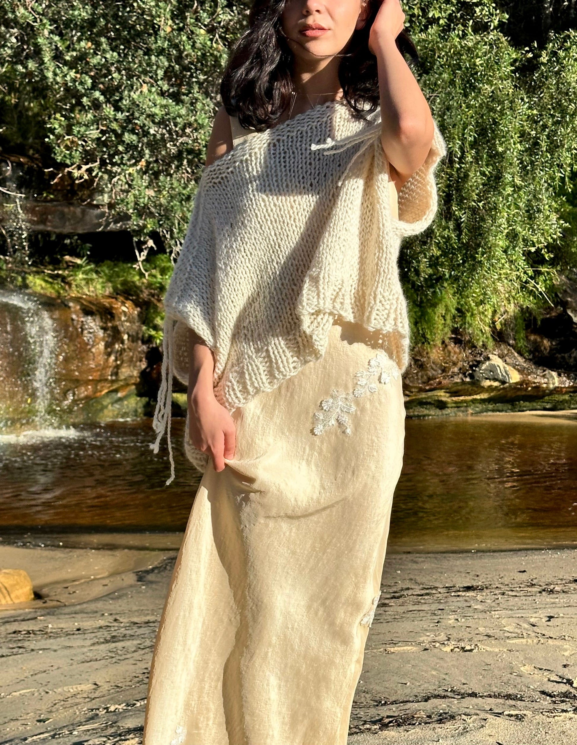 Sahara knitted wool vest, oversized cream sweater vest, sleeveless knitted vest in cream on model wearing eleanor silk dress standing up at beach waterfall