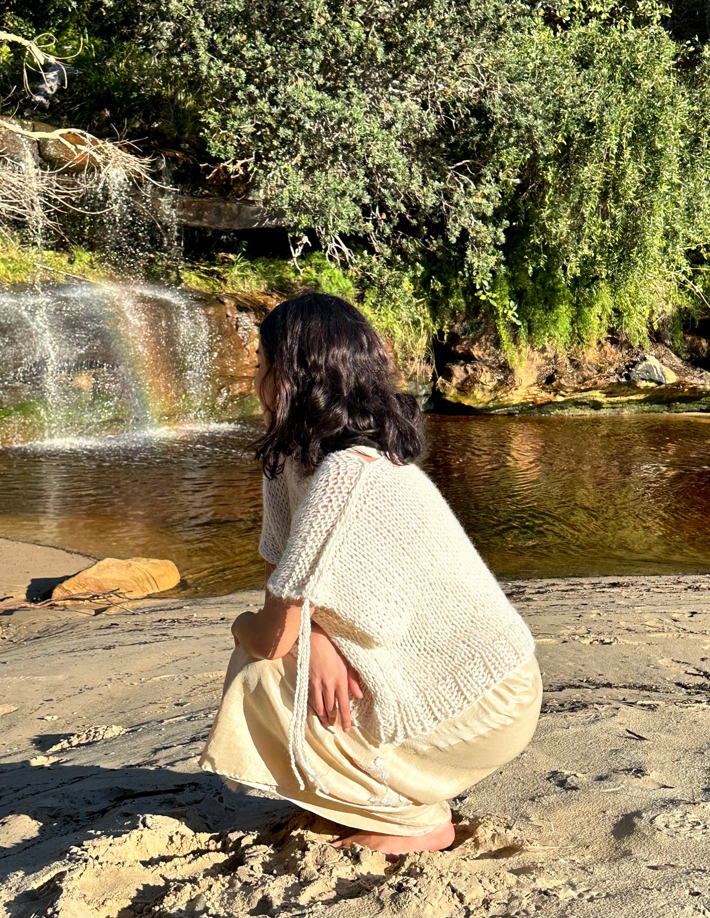 Sahara knitted wool vest, oversized cream sweater vest, sleeveless knitted vest in cream on model wearing eleanor silk dress squatting at beach waterfall